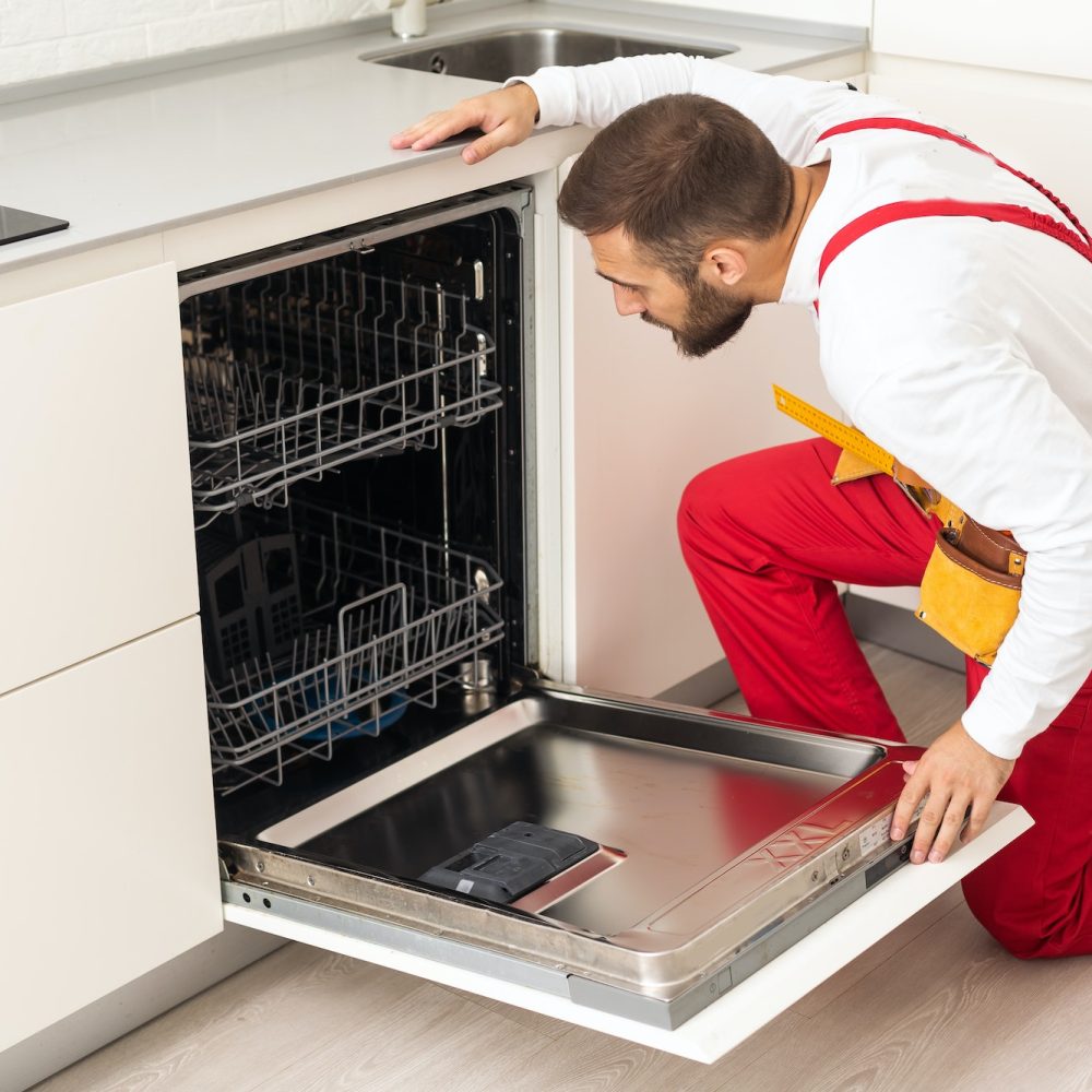 Young modern serviceman in worker suit during the repairing of the dishwasher on the domestic